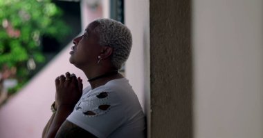 Derin düşüncelere sahip bir kadın, duvara yaslanmış, elleri birbirine kenetlenmiş dua, duygusal rahatlama, Afrika kökenli, yansıtıcı an, açık hava