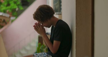 Duygusal sıkıntı içindeki genç bir çocuk, duvara yaslanmış, elleri yüzünü kapatmış, Afrika kökenli, açık hava manzaralı, kederli bir ifade, kişisel mücadele