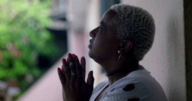 Umut dolu bir kadın, duvara yaslanmış, elleri dua ederken kenetlenmiş, Afrika kökenli, açık hava manzaralı, iyimser bir yüz ifadesi olan.