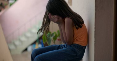 Küçük çocuk elleriyle yüzünü kapatıyor, duygusal çöküntü içinde yerde oturuyor, huzurlu ama kasvetli bir şehir ortamını yansıtıyor, üzüntü ve travma yaşıyor.