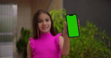 Genç kız kameraya yakın yeşil ekran krom anahtarı ile akıllı telefonu tutuyor, sıcak bir şekilde gülümsüyor, açık bir evde kişiselleştirilebilir içerik için boş ekranı gösteriyor