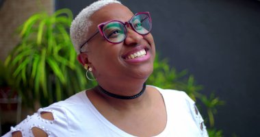 Afro-Amerikalı bir kadın, parlak bir gülümsemeyle, düşünceli ve neşeli bir ifadeyle, yeşillik ve kendine güvenle dışarı bakıyor.