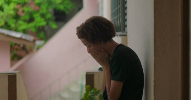 Duygusal sıkıntı içindeki genç bir çocuk, duvara yaslanmış, elleri yüzünü kapatmış, Afrika kökenli, açık hava manzaralı, kederli bir ifade, kişisel mücadele