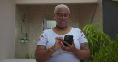 Akıllı telefon kullanan Afrika kökenli bir kadın, odaklanmış ve meşgul, arka planda bitkiler ve günlük dekor ile açık bir konut ortamında