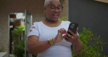 Afrika kökenli bir kadın akıllı telefon ekranına heyecanla tepki veriyor, kameraya neşeyle gösteriyor, yeşil ekran ve krom anahtar açık alanda gösteriliyor.