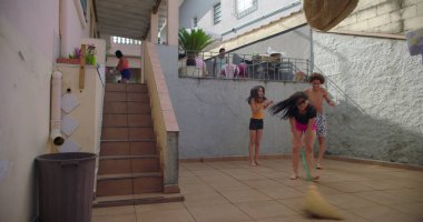 Avluda süpürgeyle oynayan çocuklar, kardeş gülüşü, kaygısız eğlence, zıplama, aile bağları, neşeli enerji, neşeli an, açık hava heyecanı. Hayatta basit şeyler.