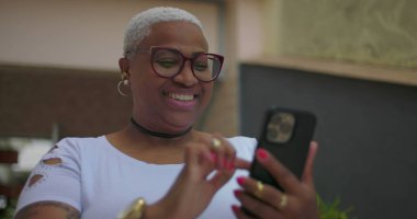 Afrika kökenli bir kadın akıllı bir telefona bakarken sevinçle tepki veriyor. Açık havada bir ev ortamında heyecan ve şaşkınlığı ifade ediyor.