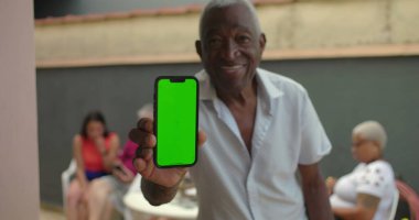 Yaşlı bir siyah adam elinde yeşil ekranlı akıllı bir telefon tutuyor, kendine güvenerek gülümsüyor, teknolojiyi açık havada sergiliyor, arka planda çeşitli yetişkinler birbiriyle etkileşime geçiyor.