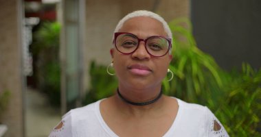 Düşünceli yüzlü, gözlüklü, kameraya bakan, özgüvenli ve arka planda yeşillik olan açık hava ortamında bestelenmiş Afrikalı Amerikalı bir kadın.