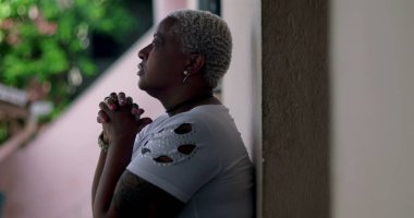 Derin düşüncelere sahip bir kadın, duvara yaslanmış, elleri birbirine kenetlenmiş dua, duygusal rahatlama, Afrika kökenli, yansıtıcı an, açık hava