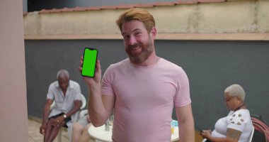 Krom tuşlu yeşil ekranlı akıllı telefonu tutan adam, kendinden emin bir şekilde gülümseyen, dijital içeriği gösteren, açık hava ayarları, teknoloji odaklı, gelişigüzel an, bağlantı ekranı