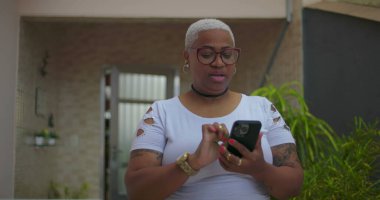 Akıllı telefon kullanan Afrika kökenli bir kadın, odaklanmış ve meşgul, arka planda bitkiler ve günlük dekor ile açık bir konut ortamında