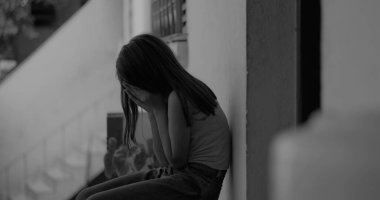 Elleri kapalı, gözleri kapalı, duygusal bir anda yerde oturan genç bir çocuk, sakin, yansıtıcı şehir ortamında travma ve üzüntü ile başa çıkıyor, siyah ve beyaz