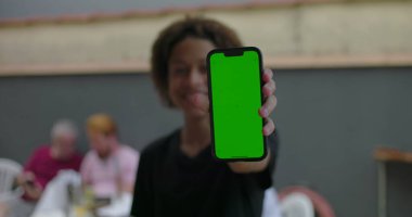 Genç, yeşil ekranlı akıllı telefonu tutuyor, kendine güvenerek gülümsüyor, çeşitli yetişkinlerin bulanık arka planda etkileşim kurduğu sıradan bir toplantıda.