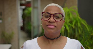 Düşünceli yüzlü, gözlüklü, kameraya bakan, özgüvenli ve arka planda yeşillik olan açık hava ortamında bestelenmiş Afrikalı Amerikalı bir kadın.