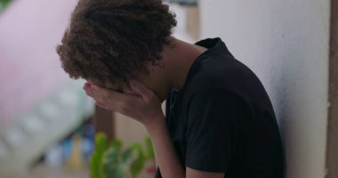Duygusal çöküntü içindeki genç çocuk, başı eğik oturuyor, elleri yüzünü kapatıyor, Afrika kökenli, bunalmış, açık havada.