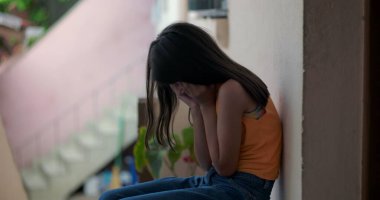 Küçük çocuk elleriyle yüzünü kapatıyor, duygusal çöküntü içinde yerde oturuyor, huzurlu ama kasvetli bir şehir ortamını yansıtıyor, üzüntü ve travma yaşıyor.