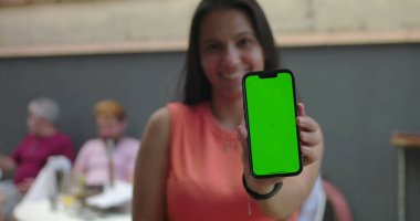 Gülümseyen ve elinde yeşil ekran ile kameraya doğru uzanan akıllı telefonu tutan bir kadın, açık havada duran, arka planda bulanık bir grup sohbeti, kroma anahtarı.