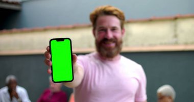 Kameraya yeşil ekranlı akıllı telefonu tutan adam, coşkuyla gülümseyen, kişiselleştirme için mobil ekran sunan, arka planda bulanık insanlar