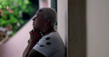 Derin düşüncelere sahip bir kadın, duvara yaslanmış, elleri birbirine kenetlenmiş dua, duygusal rahatlama, Afrika kökenli, yansıtıcı an, açık hava