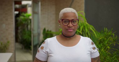 Düşünceli yüzlü, gözlüklü, kameraya bakan, özgüvenli ve arka planda yeşillik olan açık hava ortamında bestelenmiş Afrikalı Amerikalı bir kadın.