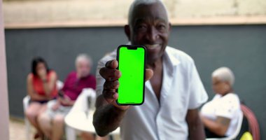 Afro-Amerikalı yaşlı bir adam elinde yeşil ekran krom anahtarıyla akıllı telefon tutuyor, avluda gülümsüyor, arka planda bulanık insanlar var.