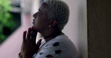 Umut dolu bir kadın, duvara yaslanmış, elleri dua ederken kenetlenmiş, Afrika kökenli, açık hava manzaralı, iyimser bir yüz ifadesi olan.