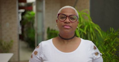 Düşünceli yüzlü, gözlüklü, kameraya bakan, özgüvenli ve arka planda yeşillik olan açık hava ortamında bestelenmiş Afrikalı Amerikalı bir kadın.