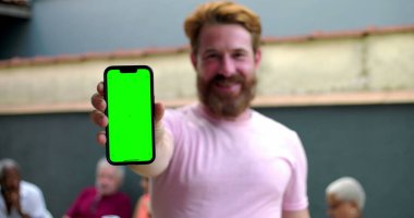 Kameraya yeşil ekranlı akıllı telefonu tutan adam, coşkuyla gülümseyen, kişiselleştirme için mobil ekran sunan, arka planda bulanık insanlar