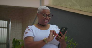 Afrika kökenli bir kadın akıllı bir telefona bakarken sevinçle tepki veriyor. Açık havada bir ev ortamında heyecan ve şaşkınlığı ifade ediyor.
