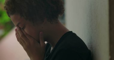 Duygusal sıkıntı içindeki genç bir çocuk, elleri yüzünü kapatıyor, ağlıyor, duvara yaslanıyor, Afrika kökenli, açık hava manzaralı, yakın plan.