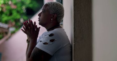 Derin düşüncelere sahip bir kadın, duvara yaslanmış, elleri birbirine kenetlenmiş dua, duygusal rahatlama, Afrika kökenli, yansıtıcı an, açık hava