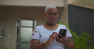 Akıllı telefon kullanan Afrika kökenli bir kadın, odaklanmış ve meşgul, arka planda bitkiler ve günlük dekor ile açık bir konut ortamında