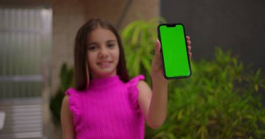Genç kız kameraya yakın yeşil ekran krom anahtarı ile akıllı telefonu tutuyor, sıcak bir şekilde gülümsüyor, açık bir evde kişiselleştirilebilir içerik için boş ekranı gösteriyor