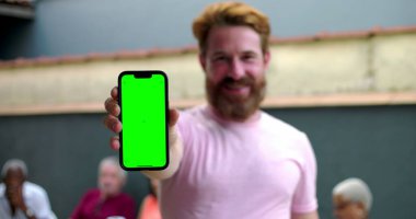 Kameraya yeşil ekranlı akıllı telefonu tutan adam, coşkuyla gülümseyen, kişiselleştirme için mobil ekran sunan, arka planda bulanık insanlar