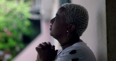 Umut dolu bir kadın, duvara yaslanmış, elleri dua ederken kenetlenmiş, Afrika kökenli, açık hava manzaralı, iyimser bir yüz ifadesi olan.