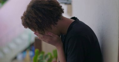 Duygusal çöküntü içindeki genç çocuk, başı eğik oturuyor, elleri yüzünü kapatıyor, Afrika kökenli, bunalmış, açık havada.