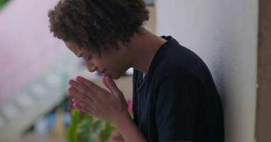 Duygusal çöküntü içindeki genç çocuk, başı eğik oturuyor, elleri yüzünü kapatıyor, Afrika kökenli, bunalmış, açık havada.