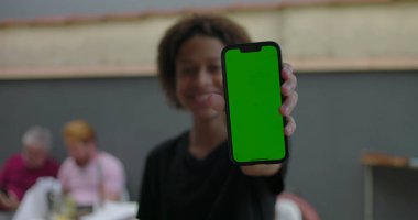 Genç, yeşil ekranlı akıllı telefonu tutuyor, kendine güvenerek gülümsüyor, çeşitli yetişkinlerin bulanık arka planda etkileşim kurduğu sıradan bir toplantıda.