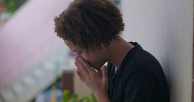 Duygusal çöküntü içindeki genç çocuk, başı eğik oturuyor, elleri yüzünü kapatıyor, Afrika kökenli, bunalmış, açık havada.