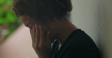 Duygusal sıkıntı içindeki genç bir çocuk, elleri yüzünü kapatıyor, ağlıyor, duvara yaslanıyor, Afrika kökenli, açık hava manzaralı, yakın plan.