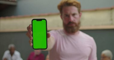 Elinde yeşil ekranlı akıllı telefon tutan adam, ciddi ifade, dışarıda duran, arka planda bulanık insanlar, kişiselleştirilebilir ekran odağı.