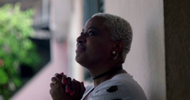 Umut dolu bir kadın, duvara yaslanmış, elleri dua ederken kenetlenmiş, Afrika kökenli, açık hava manzaralı, iyimser bir yüz ifadesi olan.