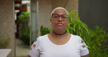 Düşünceli yüzlü, gözlüklü, kameraya bakan, özgüvenli ve arka planda yeşillik olan açık hava ortamında bestelenmiş Afrikalı Amerikalı bir kadın.