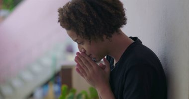 Duygusal çöküntü içindeki genç çocuk, başı eğik oturuyor, elleri yüzünü kapatıyor, Afrika kökenli, bunalmış, açık havada.