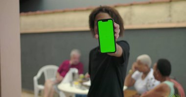 Afrika kökenli, yeşil ekranlı, krom anahtarlı, arka planda gelişigüzel sohbetlerin keyfini çıkaran çeşitli yetişkinlerin bulunduğu bir sosyal ortamda sergilenen akıllı telefon.