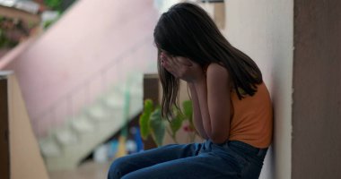 Küçük çocuk elleriyle yüzünü kapatıyor, duygusal çöküntü içinde yerde oturuyor, huzurlu ama kasvetli bir şehir ortamını yansıtıyor, üzüntü ve travma yaşıyor.