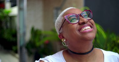 Afro-Amerikalı bir kadın, parlak bir gülümsemeyle, düşünceli ve neşeli bir ifadeyle, yeşillik ve kendine güvenle dışarı bakıyor.