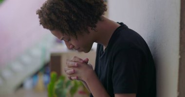 Duygusal çöküntü içindeki genç çocuk, başı eğik oturuyor, elleri yüzünü kapatıyor, Afrika kökenli, bunalmış, açık havada.