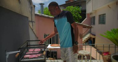 Afrika kökenli Amerikalı bir adam çatıdaki barbeküde et pişiriyor, biftek ve sosisleri kontrol ediyor ve canlı bir şehir ortamında yemek hazırlıyor.